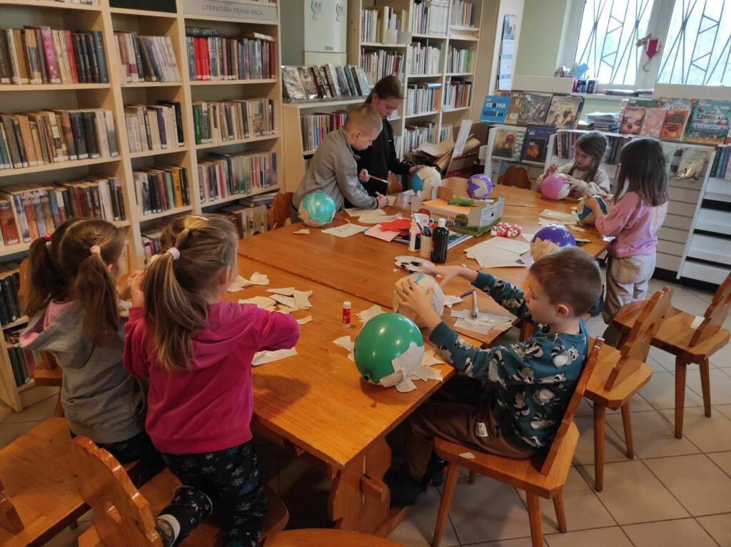 na zdjęciu widać salę biblioteczną , na środku pomieszczenia stoi stół, przy stole siedzą dzieci , wykonują pracę plastyczną