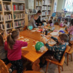 na zdjęciu widać salę biblioteczną , na środku pomieszczenia stoi stół, przy stole siedzą dzieci , wykonują pracę plastyczną
