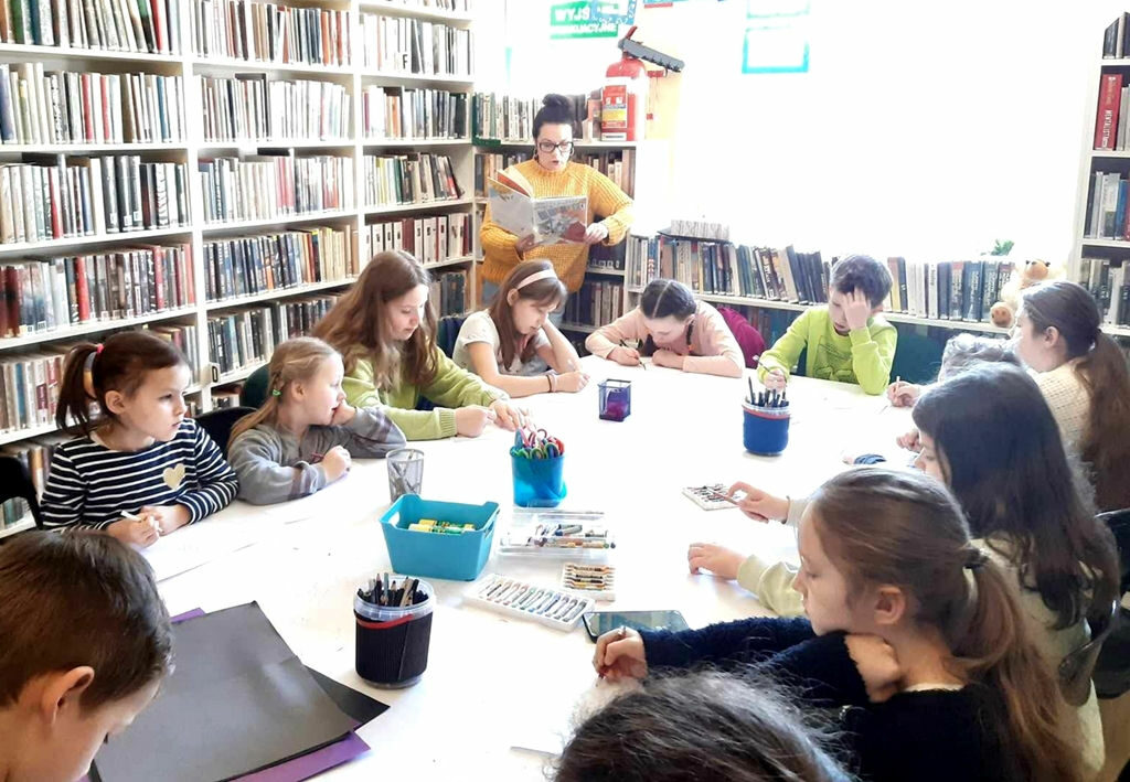na zdjęciu widać dzieci siedzące wokół stołu w bibliotece i czytająca im kobietę