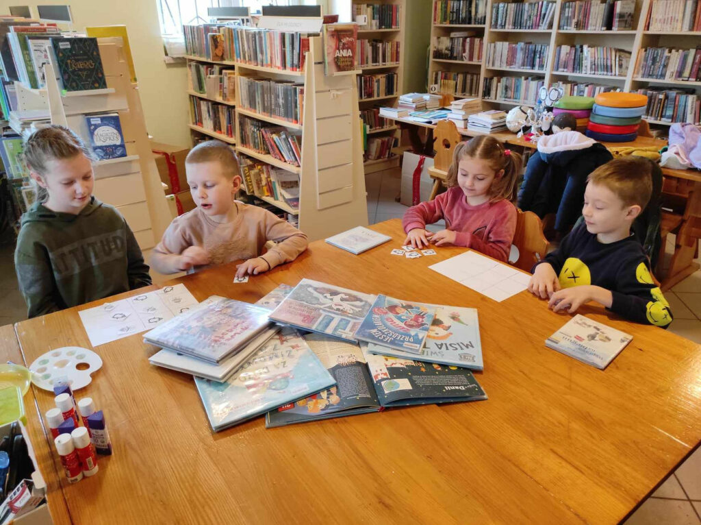 na zdjęciu widać pomieszczenie biblioteczne, przy stole siedzą dzieci, biorą udział w zabawie