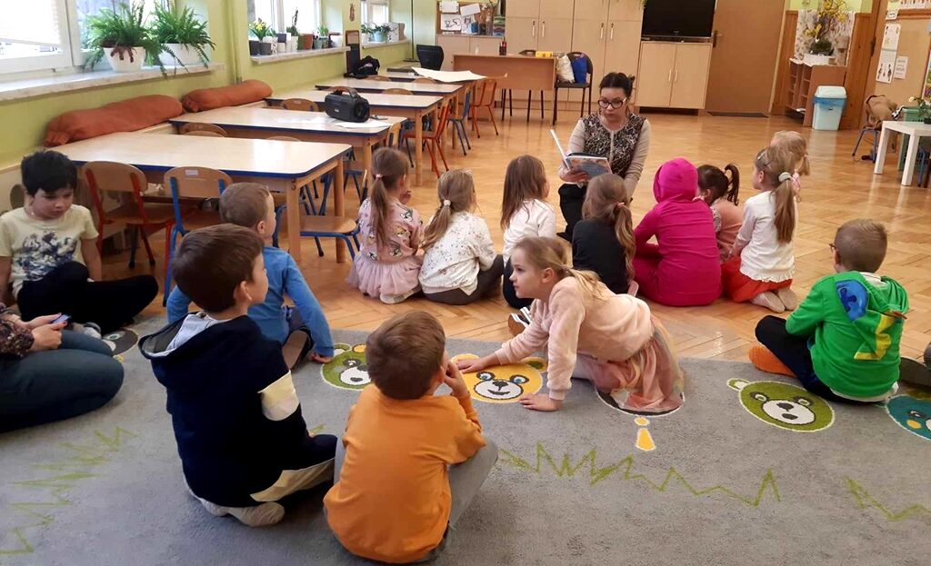 na zdjęciu widać dzieci siedzące na dywanie w sali przedszkolnej słuchające czytanej im książki