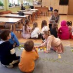 na zdjęciu widać dzieci siedzące na dywanie w sali przedszkolnej słuchające czytanej im książki