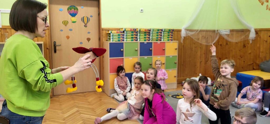na zdjęciu widać dzieci siedzące na dywanie i pokazującą im maskotkę kobietę