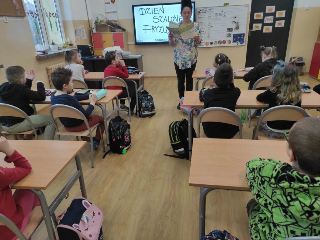 na zdjęciu widać salę szkolną, dzieci siedzą przy stolikach, bibliotekarz czyta opowiadanie