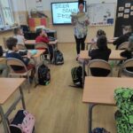 na zdjęciu widać salę szkolną, dzieci siedzą przy stolikach, bibliotekarz czyta opowiadanie