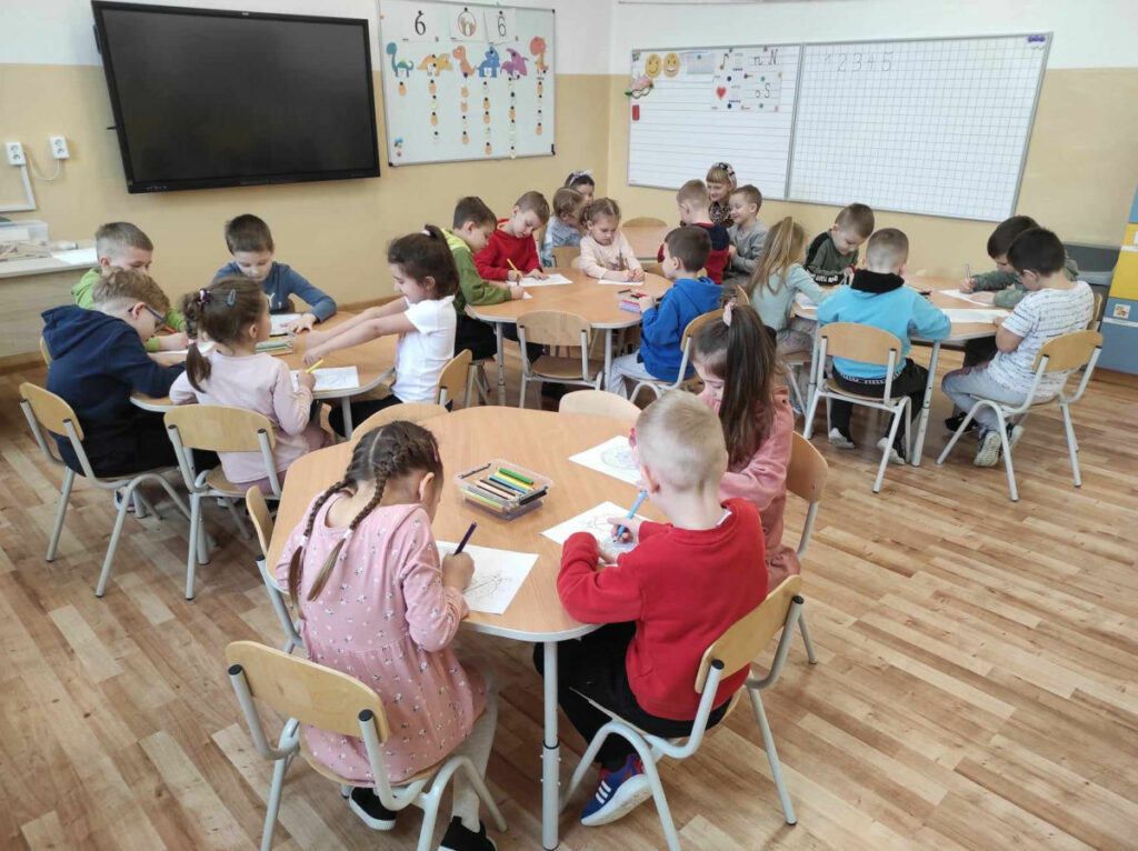 na zdjęciu widać salę przedszkolną, dzieci siedzą przy stolikach, wykonują pracę plastyczną