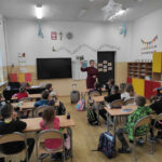 na zdjęciu widać salę szkolną, dzieci siedzą przy stolikach, słuchają książki czytanej przez bibliotekarza