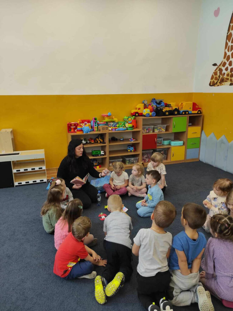 na zdjęciu widać salę przedszkolną, dzieci siedzą w kole na dywanie, bibliotekarz czyta książkę oraz pokazuje elementy opowiadania