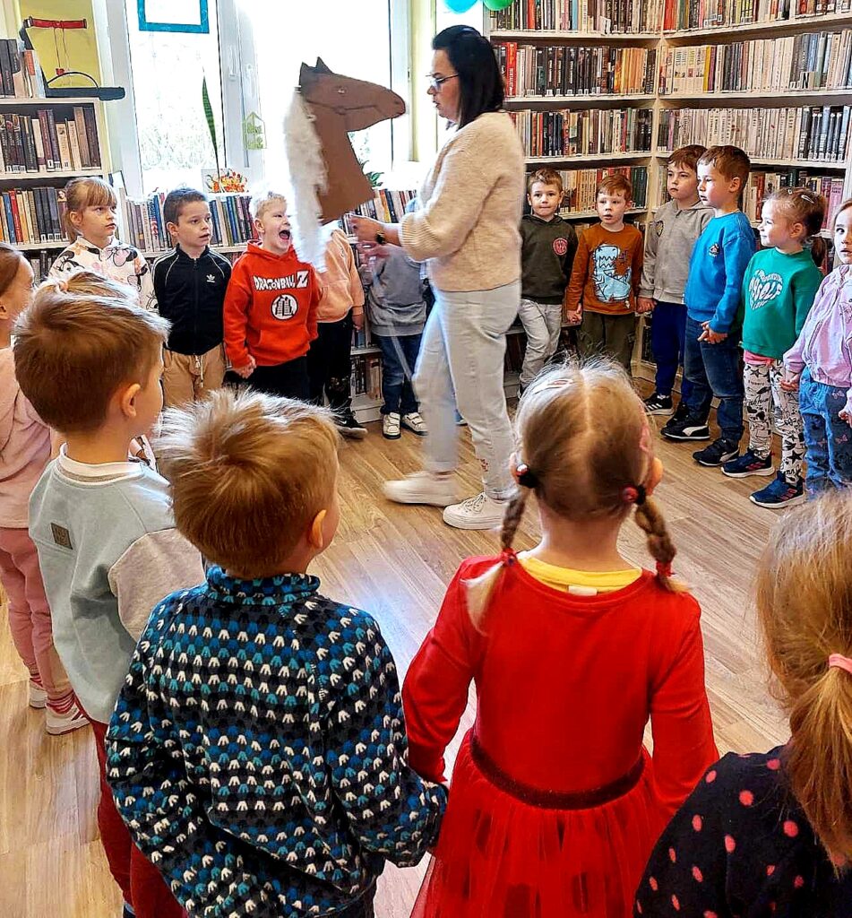 na zdjęciu widać dzieci stojące w kole oraz bibliotekarkę trzymającą papierowego konia