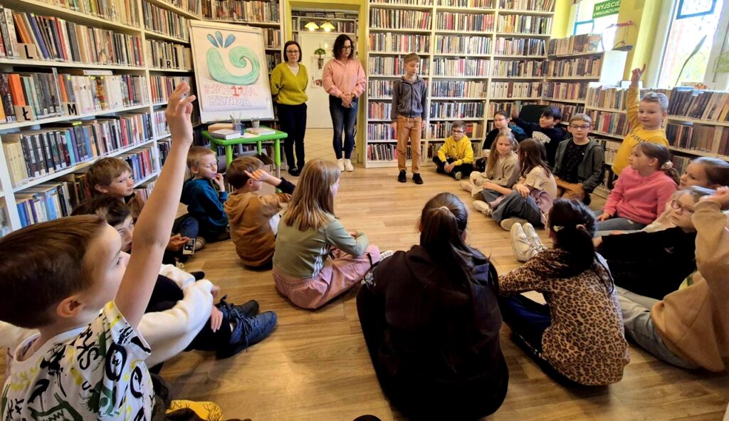 na zdjęciu widać dzieci i dwie bibliotekarki w sali bibliotecznej