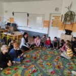 na zdjęciu widać salę szkolną, uczniowie siedzą w kole na dywanie, bibliotekarz czyta książkę