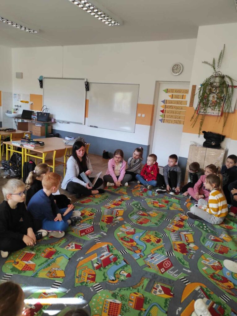 na zdjęciu widać salę szkolną, uczniowie siedzą w kole na dywanie, bibliotekarz czyta książkę
