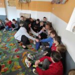 na zdjęciu widać salę szkolną, uczniowie siedzą w kole na dywanie i biorą udział w zabawie