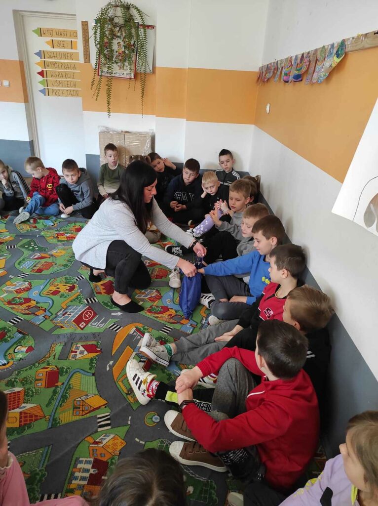 na zdjęciu widać salę szkolną, uczniowie siedzą w kole na dywanie i biorą udział w zabawie