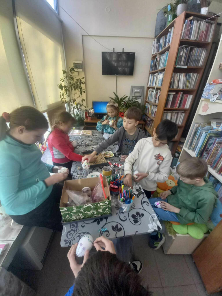 Siedmioro dzieci siedzi przy stole w bibliotece i ozdabiaja jajka wielkanocne. Na stole materiały plastyczne.