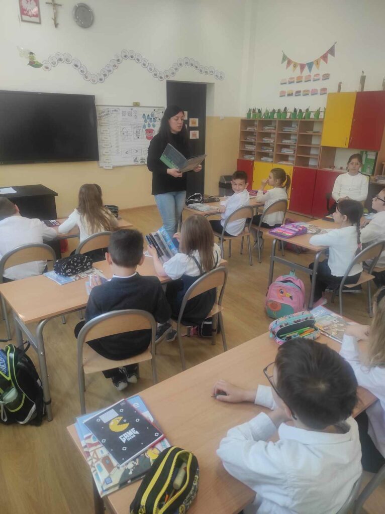 na zdjęciu widać sale szkolną, dzieci siedzą przy stolikach, bibliotekarz czyta książkę