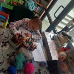 Przy stole siedzi dziewczynka i koloruje kolorowankę. Na stole flamastry, włóczki.