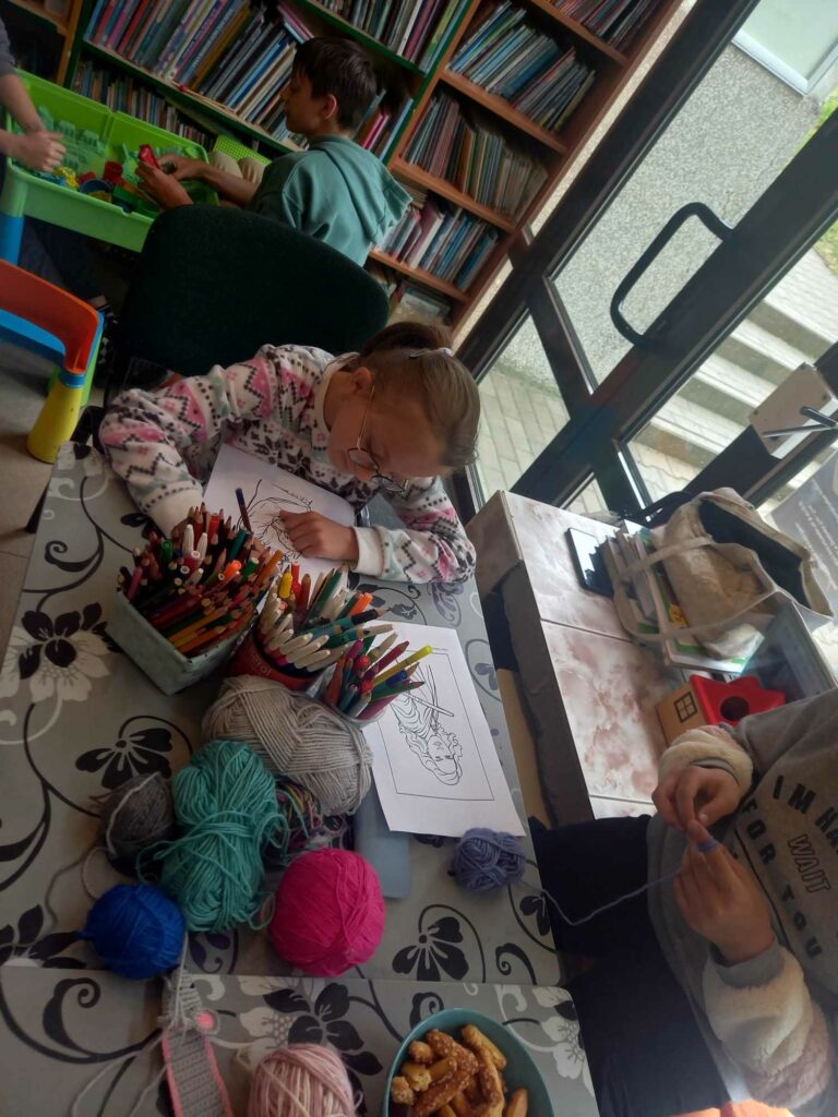 Przy stole siedzi dziewczynka i koloruje kolorowankę. Na stole flamastry, włóczki.