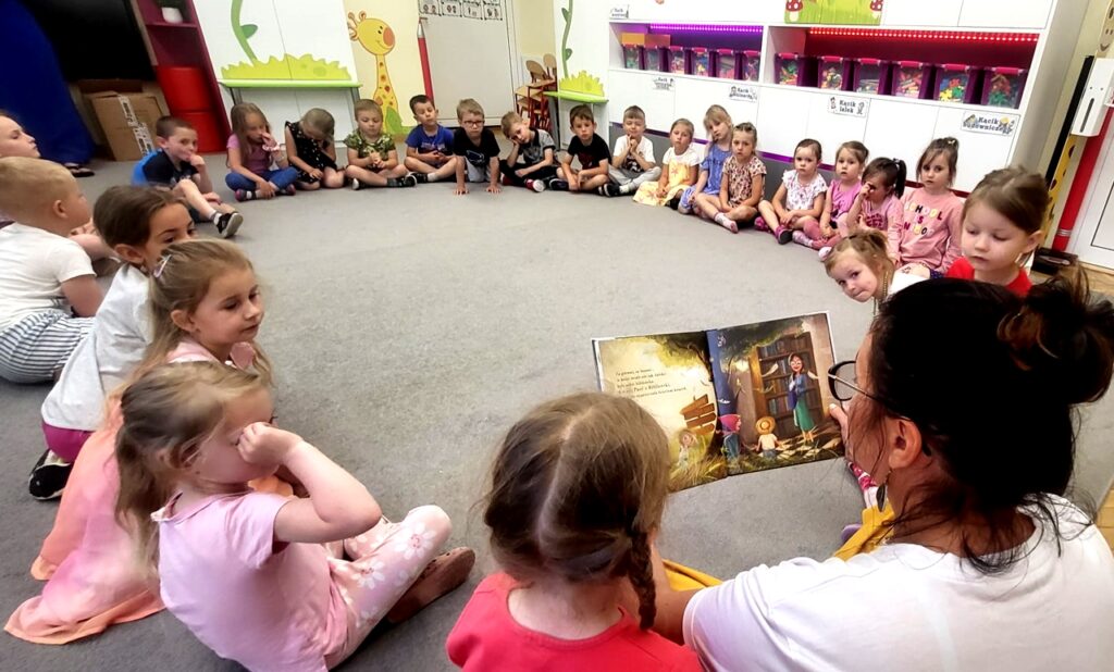 na zdjęciu widać dzieci siedzące na podłodze i słuchające czytanej im książki