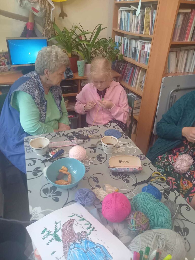 Przy stole dziedzi dziewczynka i pani. Starsza pani uczy dziewczynkę robić na szydełku. Na stole kolorowe włóczki.