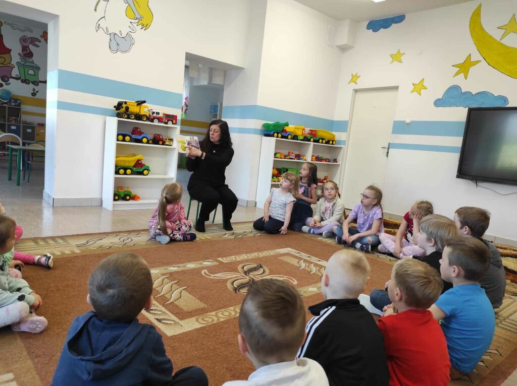 na zdjęciu widać salę przedszkolną, dzieci siedzą w kole na dywanie, bibliotekarz czyta książkę