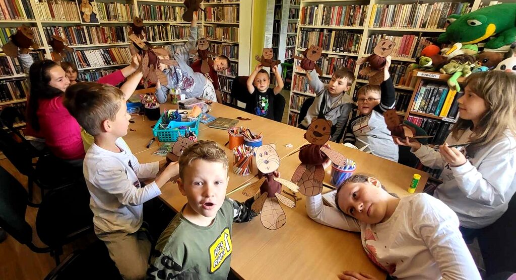 Na zdjęciu widać siedzące przy stole dzieci w sali bibliotecznej. Dzieci trzymają wykonane przez siebie bobry.