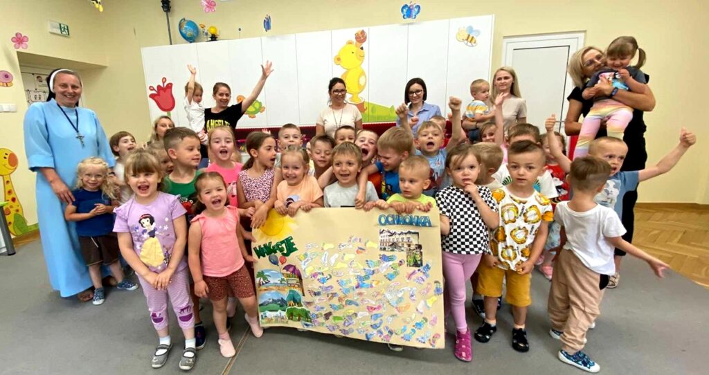 na zdjęciu widać dzieci w sali przedszkolnej wraz z Paniami. Dzieci trzymają kolorowy plakat.