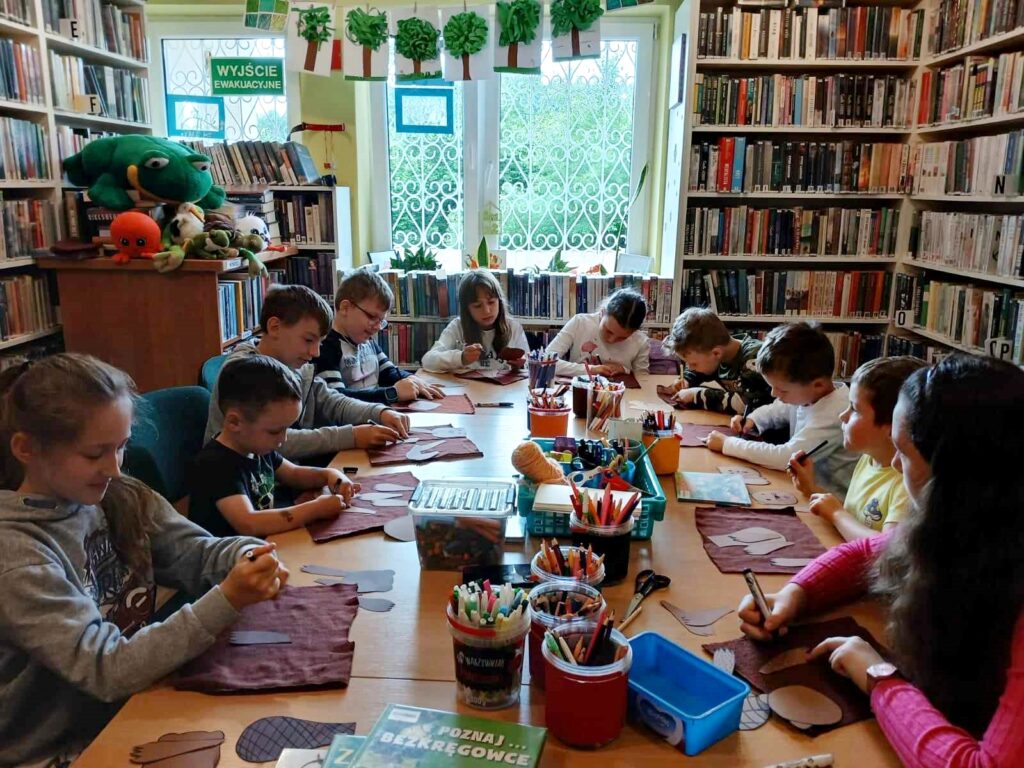 Na zdjęciu widać siedzące przy stole dzieci w sali bibliotecznej. Dzieci rysują.