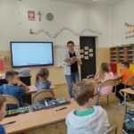 na zdjęciu widać salę szkolną, dzieci siedzą przy stolikach, bibliotekarz prowadzi zabawę kalambury