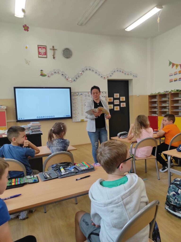 na zdjęciu widać salę szkolną, dzieci siedzą przy stolikach, bibliotekarz prowadzi zabawę kalambury