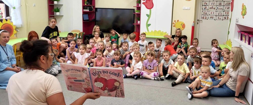 na zdjęciu widać dzieci siedzące na podłodze i słuchające czytanej im książki