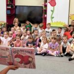 na zdjęciu widać dzieci siedzące na podłodze i słuchające czytanej im książki