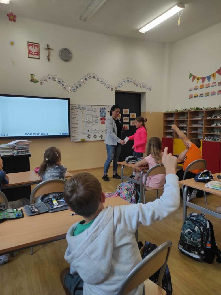 na zdjęciu widać salę szkolną, dzieci siedzą przy stolikach, bibliotekarz prowadzi zabawę kalambury
