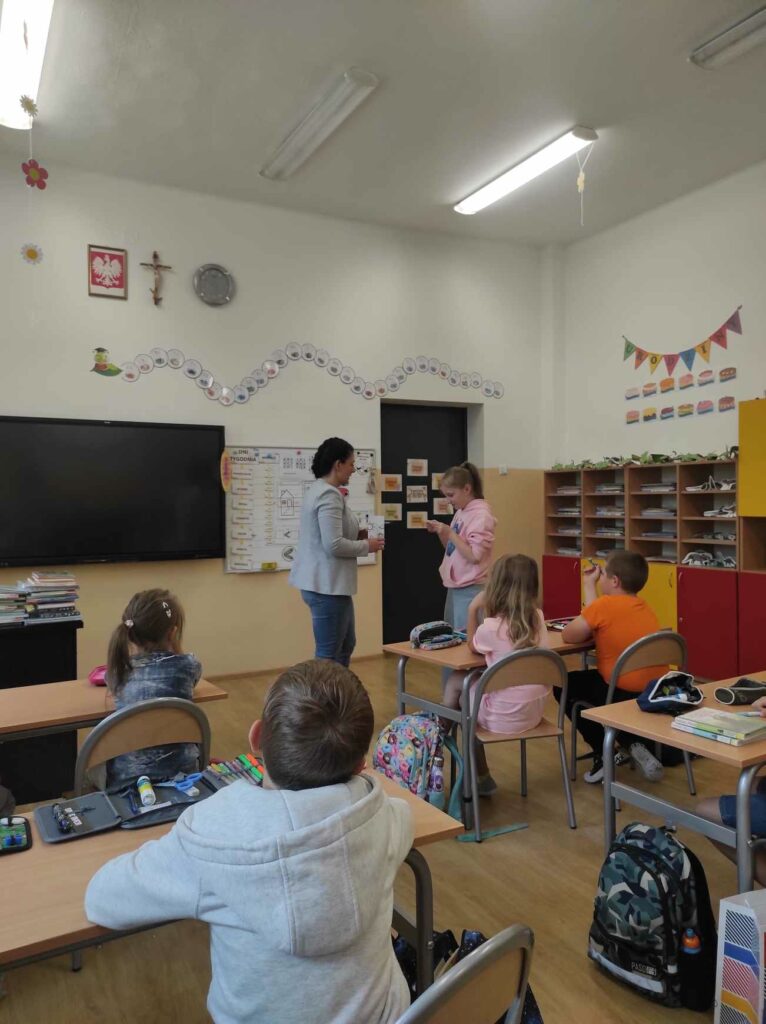 na zdjęciu widać salę szkolną, dzieci siedzą przy stolikach, bibliotekarz prowadzi zabawę kalambury