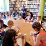 na zdjęciu widać dzieci siedzące wokół stołu oraz czytającą im bibliotekarkę