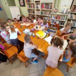 na zdjęciu widać salę biblioteczną, wokół stołu siedzą dzieci, wykonują pracę plastyczną