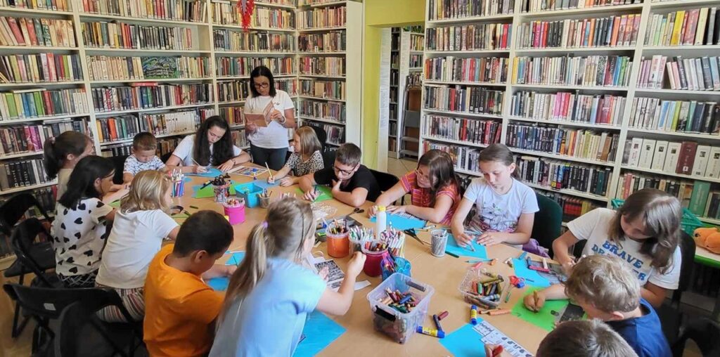 na zdjęciu widać dzieci siedzące przy stole w sali bibliotecznej i czytającą im bibliotekarkę