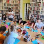 na zdjęciu widać dzieci siedzące przy stole w sali bibliotecznej i czytającą im bibliotekarkę