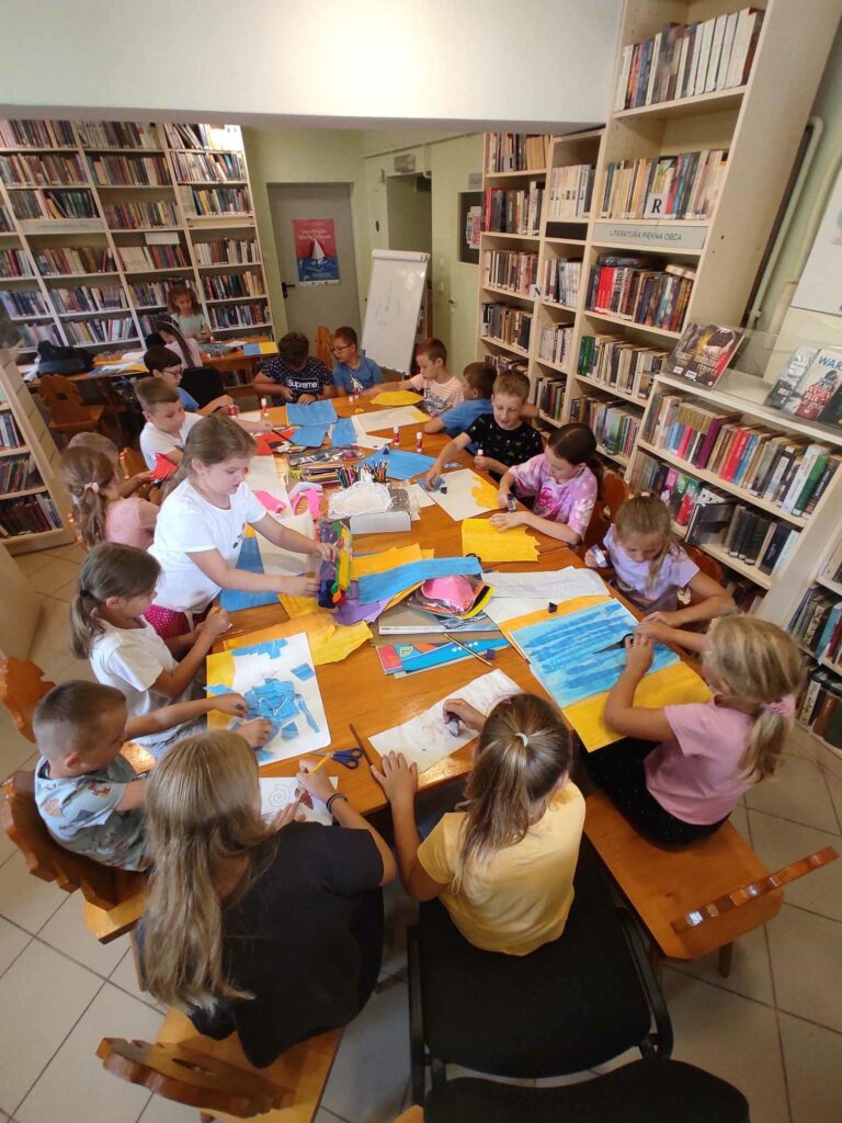 na zdjęciu widać salę biblioteczną, dzieci siedzą przy stole, wykonują pracę plastyczną
