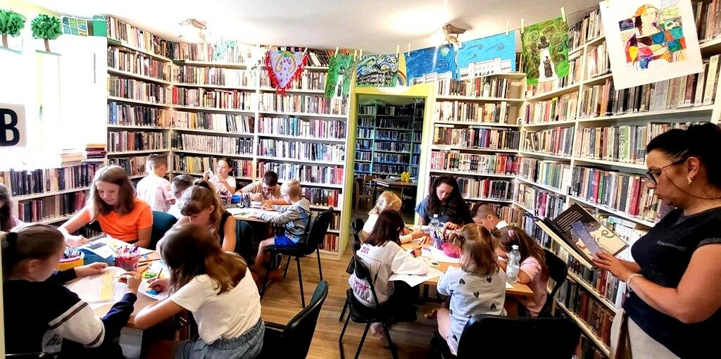 widać dzieci trzymające prace plastyczne, stoją one na tle regału z ksiązkami. Przy stołach siedzą dzieci. Pani bibliotekarka stoi i czyta książkę.