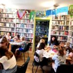 widać dzieci trzymające prace plastyczne, stoją one na tle regału z ksiązkami. Przy stołach siedzą dzieci. Pani bibliotekarka stoi i czyta książkę.