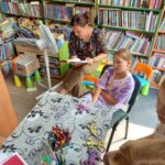 Na zdjęciu sala biblioteczna a wniej siedzi bibliotekarka i czyta bajkę. Przystole siedzą dzieci i słuchają