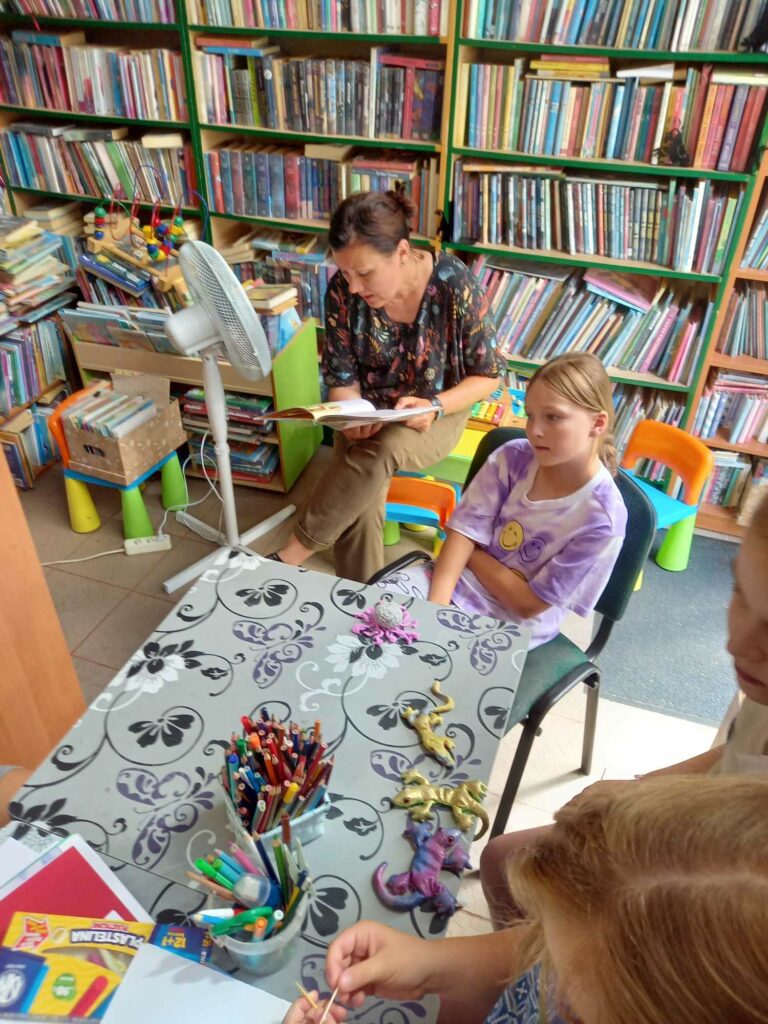Na zdjęciu sala biblioteczna a wniej siedzi bibliotekarka i czyta bajkę. Przystole siedzą dzieci i słuchają