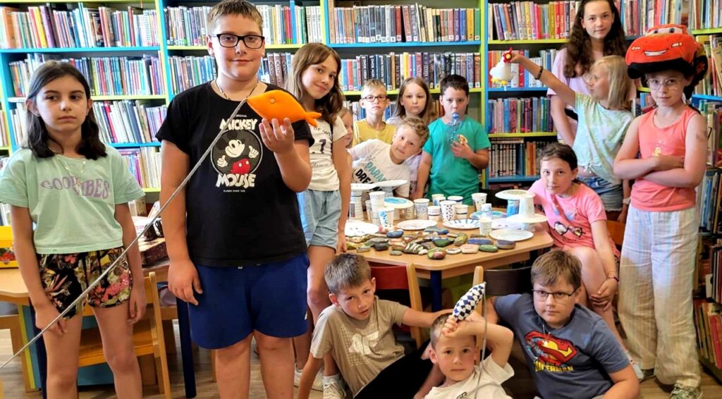 na zdjęciu widać dzieci stojące w sali bibliotecznej na tle regałów z książkami.