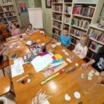 na zdjęciu widać salę biblioteczną, przy stole siedzą dzieci, wykonują prace z gliny