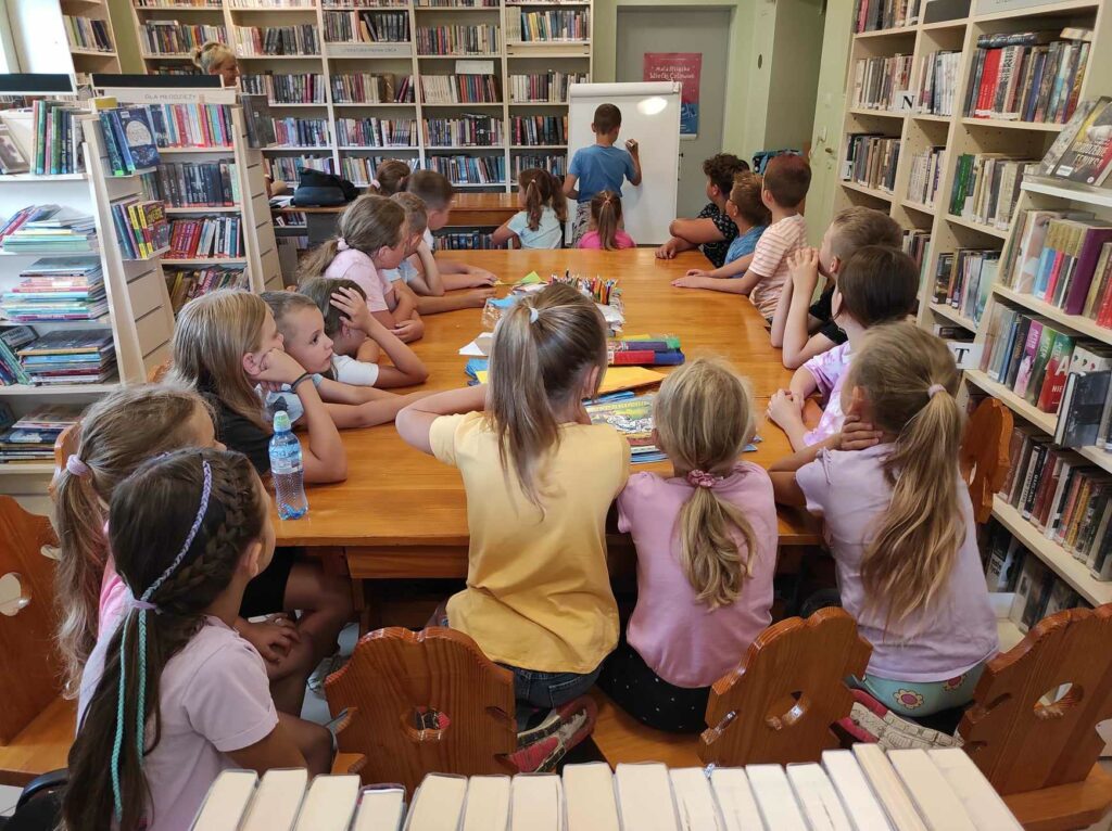 na zdjęciu widać salę biblioteczną, dzieci siedzą przy stole, bawią się w kalambury