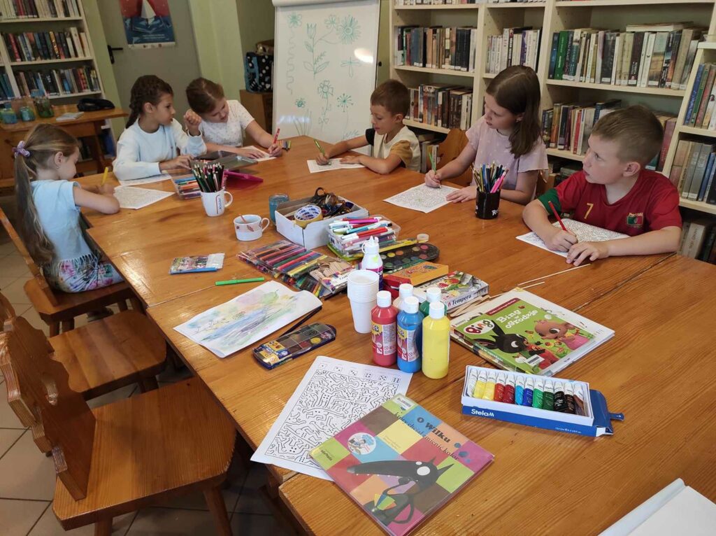 na zdjęciu widać salę biblioteczną, dzieci siedzą przy stole, po wysłuchaniu książki wykonują zadanie