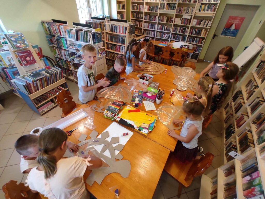 na zdjęciu widać salę biblioteczną, przy stole siedzą dzieci, projektują witraże