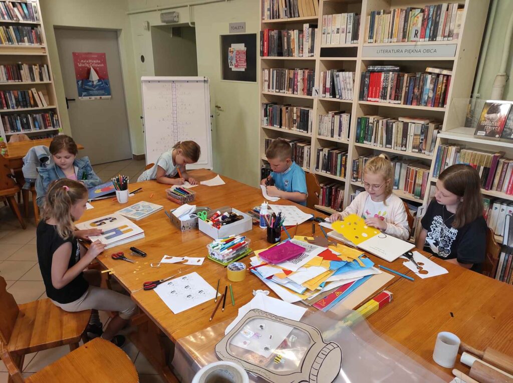 na zdjęciu widać salę biblioteczną, przy stole siedzą dzieci, przeglądają książki wybrane przez bibliotekarza