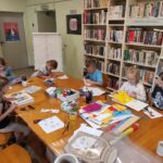 na zdjęciu widać salę biblioteczną, przy stole siedzą dzieci, przeglądają książki wybrane przez bibliotekarza